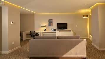 Habitación de hotel con sofá color beige, un escritorio con silla, televisor de pantalla plana, lámpara y alfombra estampada.