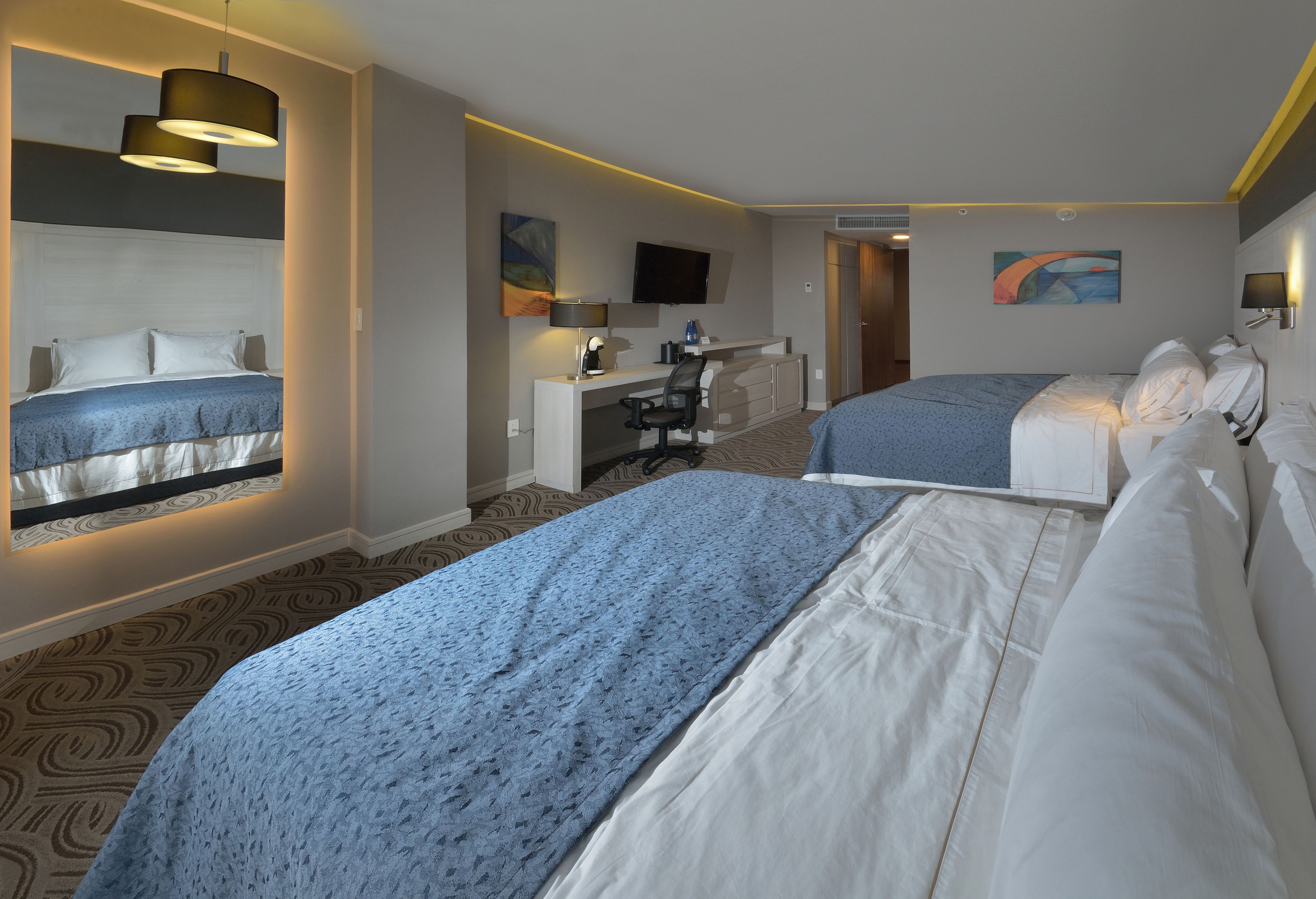 A hotel room with two beds, a desk, a chair, a TV, and artwork on the walls.