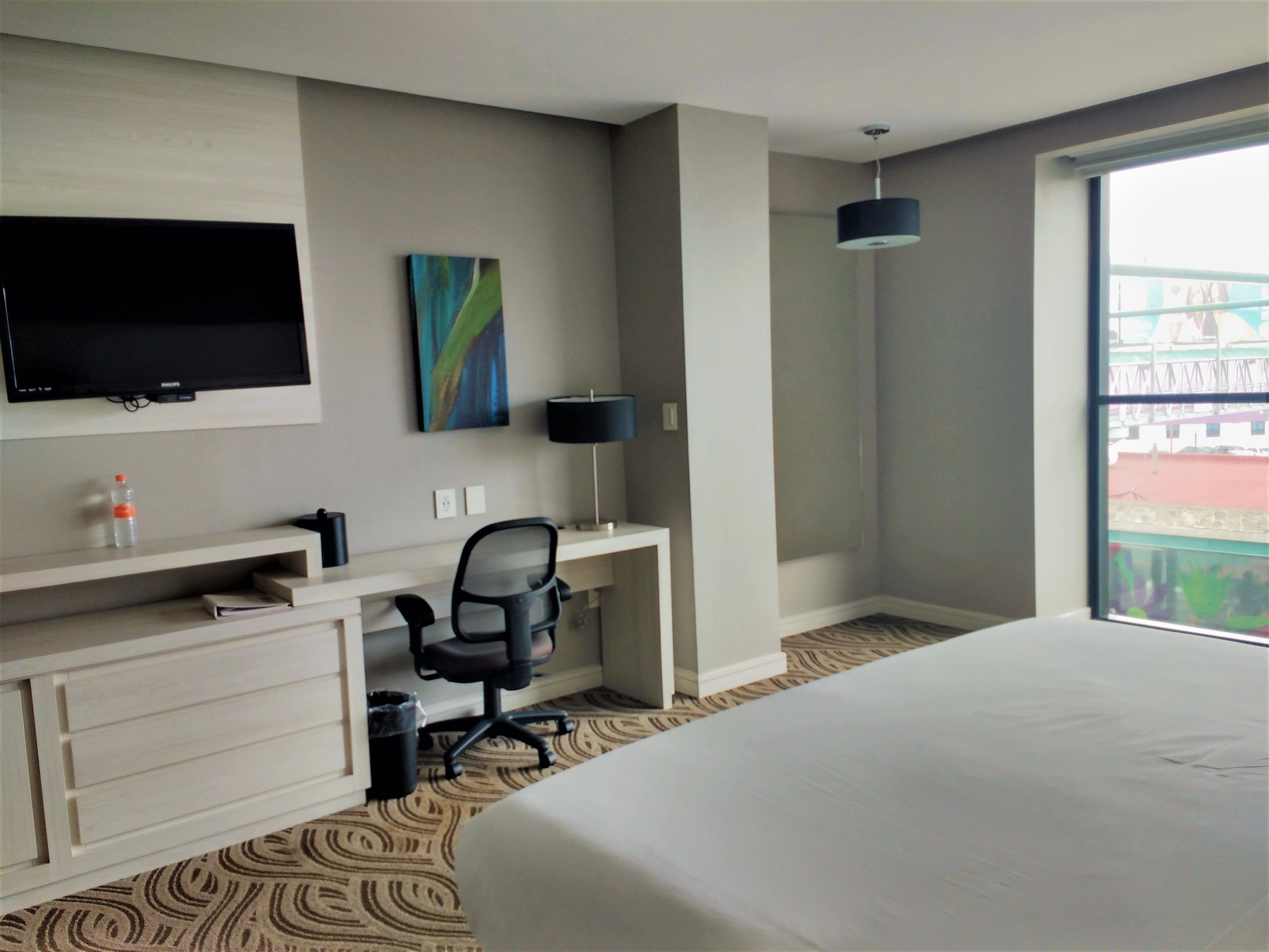 Habitación de hotel con cama, escritorio con silla, televisor, un cuadro y ventana con vistas.