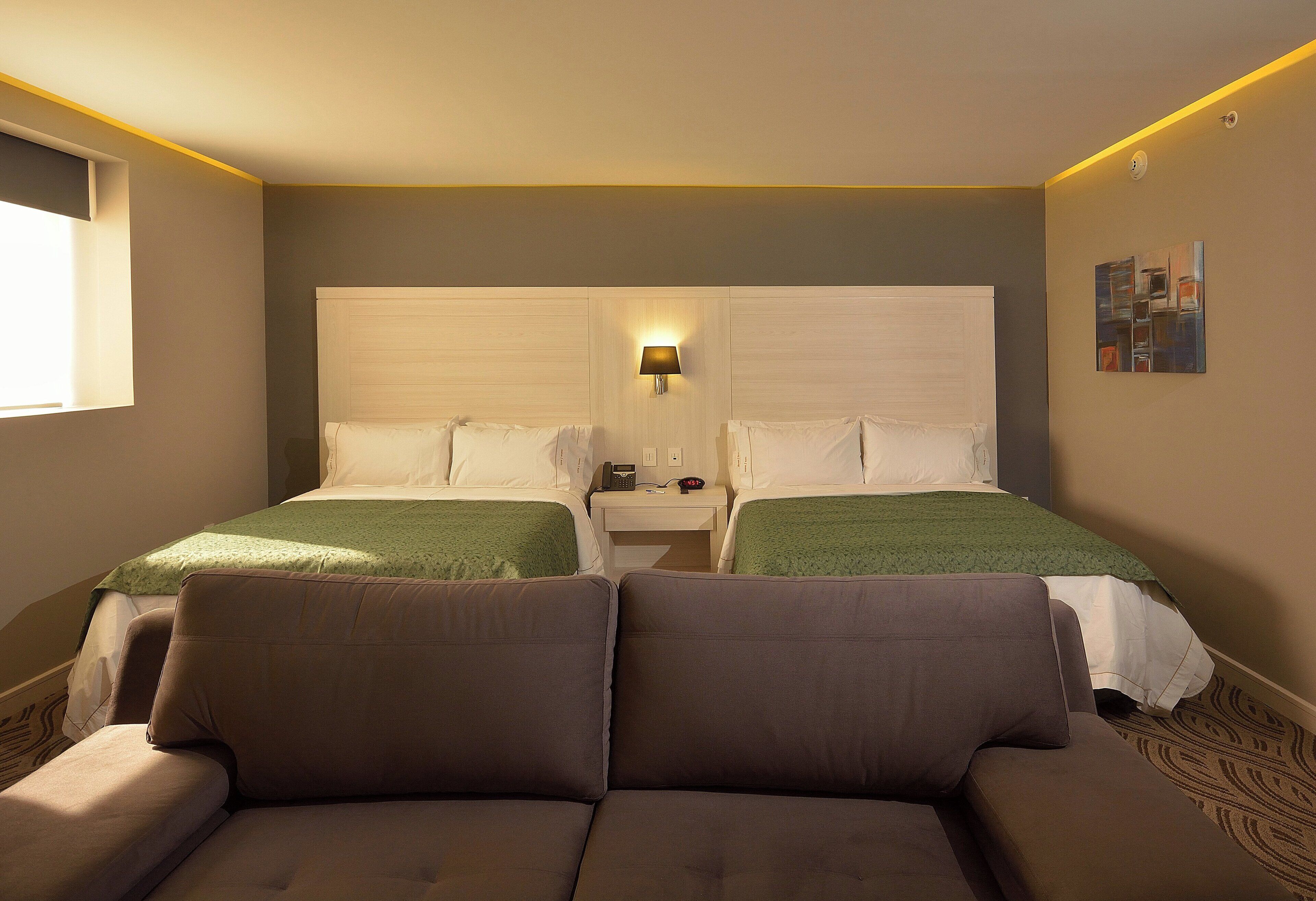 Una habitación de hotel con dos camas, un sofá, una mesita de noche con lámpara y un cuadro en la pared.