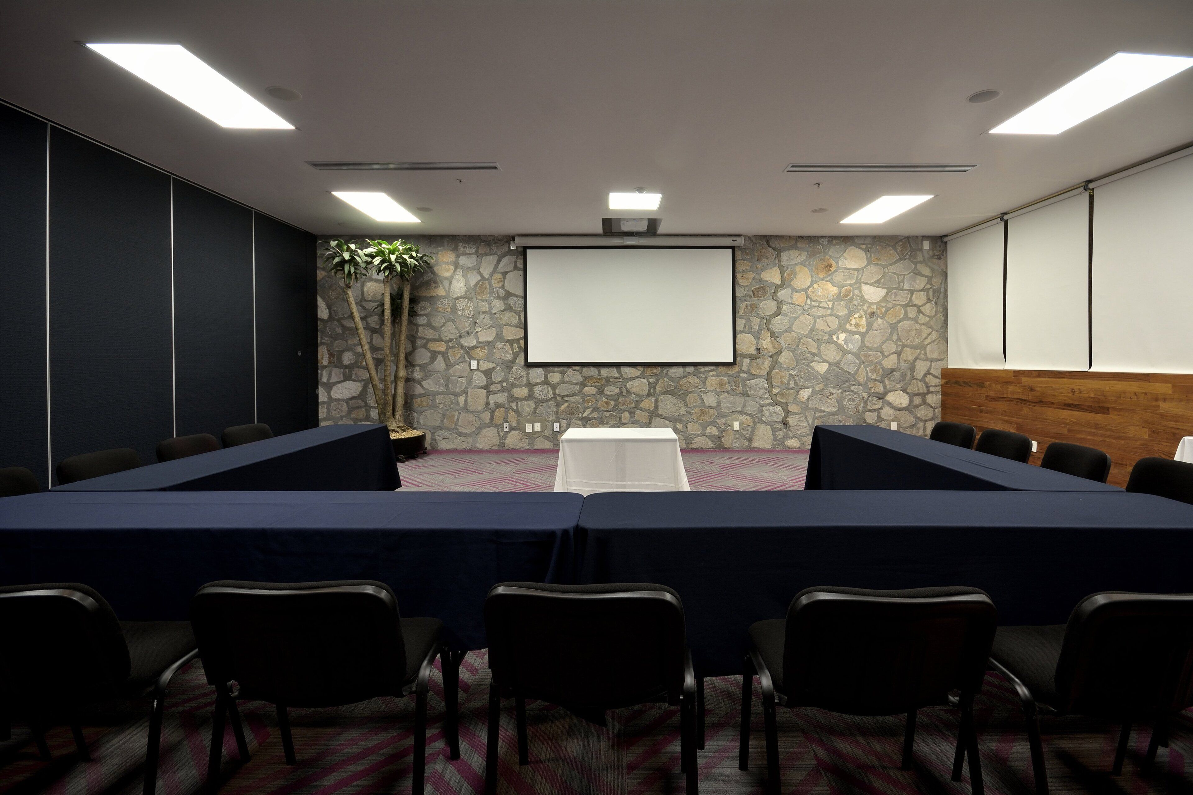 A conference room with a long table, chairs, a projector screen, and a stone wall.