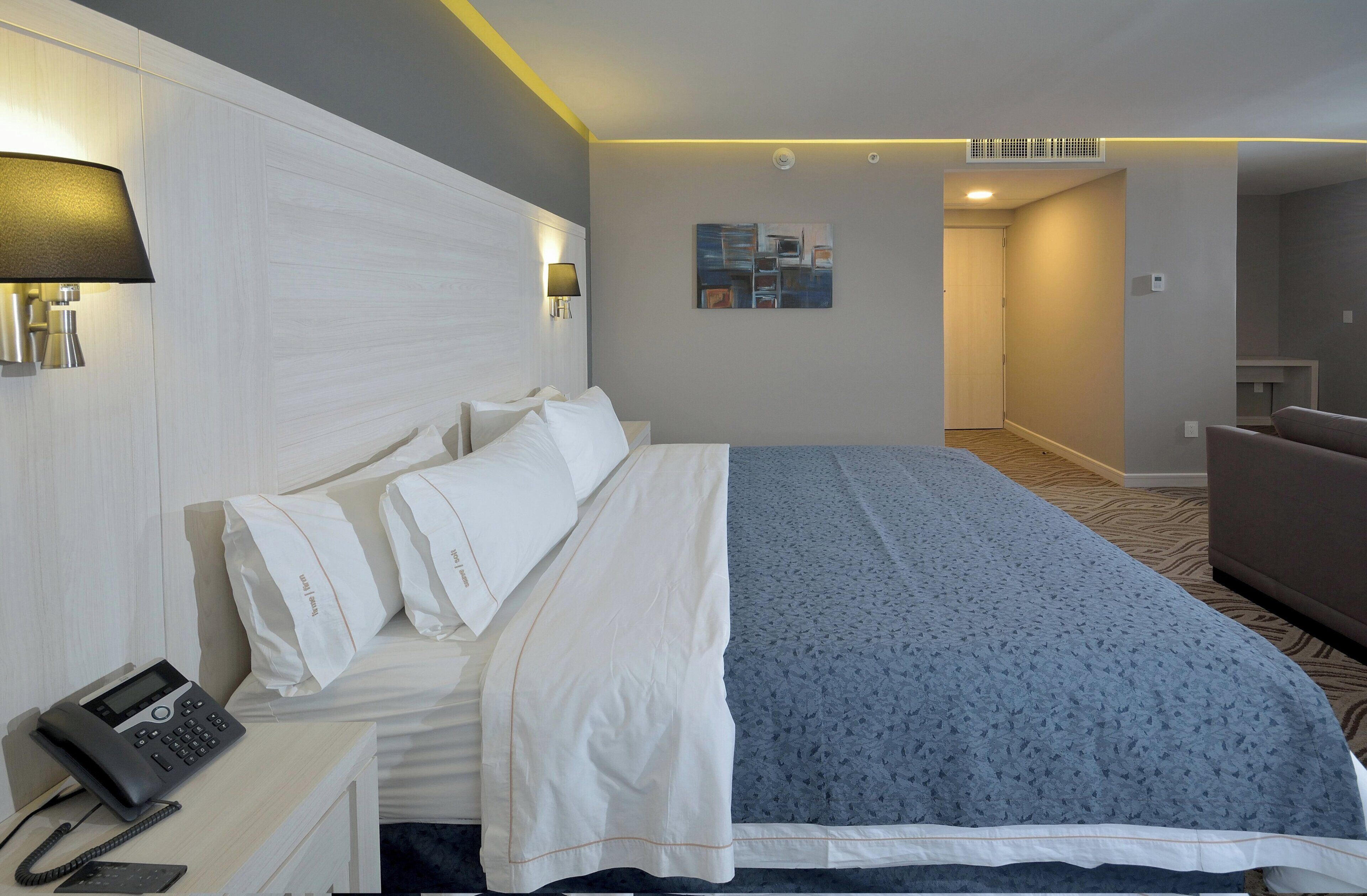 Habitación de hotel con cama, mesita de noche con teléfono, lámpara, un cuadro en la pared y puerta que da a otra habitación.