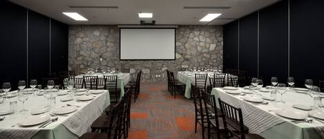 A conference room with tables set for dining, a projector screen, and a stone wall.