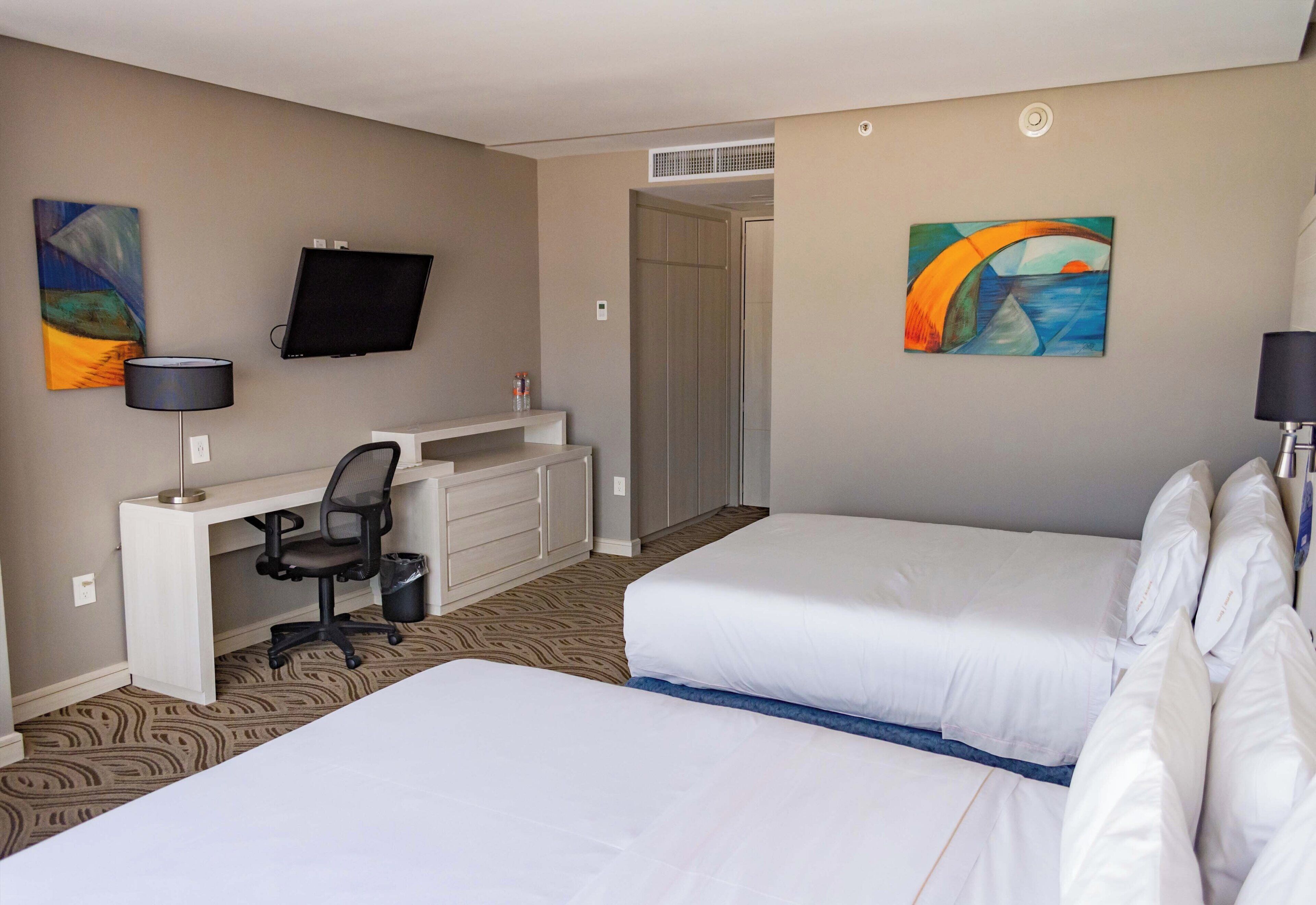 Habitación de hotel con dos camas, un escritorio con silla, televisión y obras de arte en las paredes.
