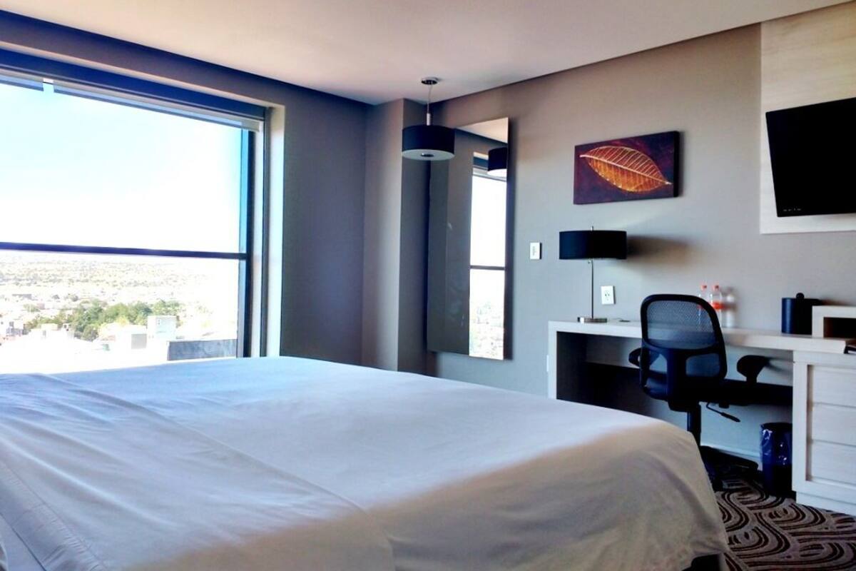 A hotel room with a large bed, a desk with a chair, a television, and a window offering a city view.
