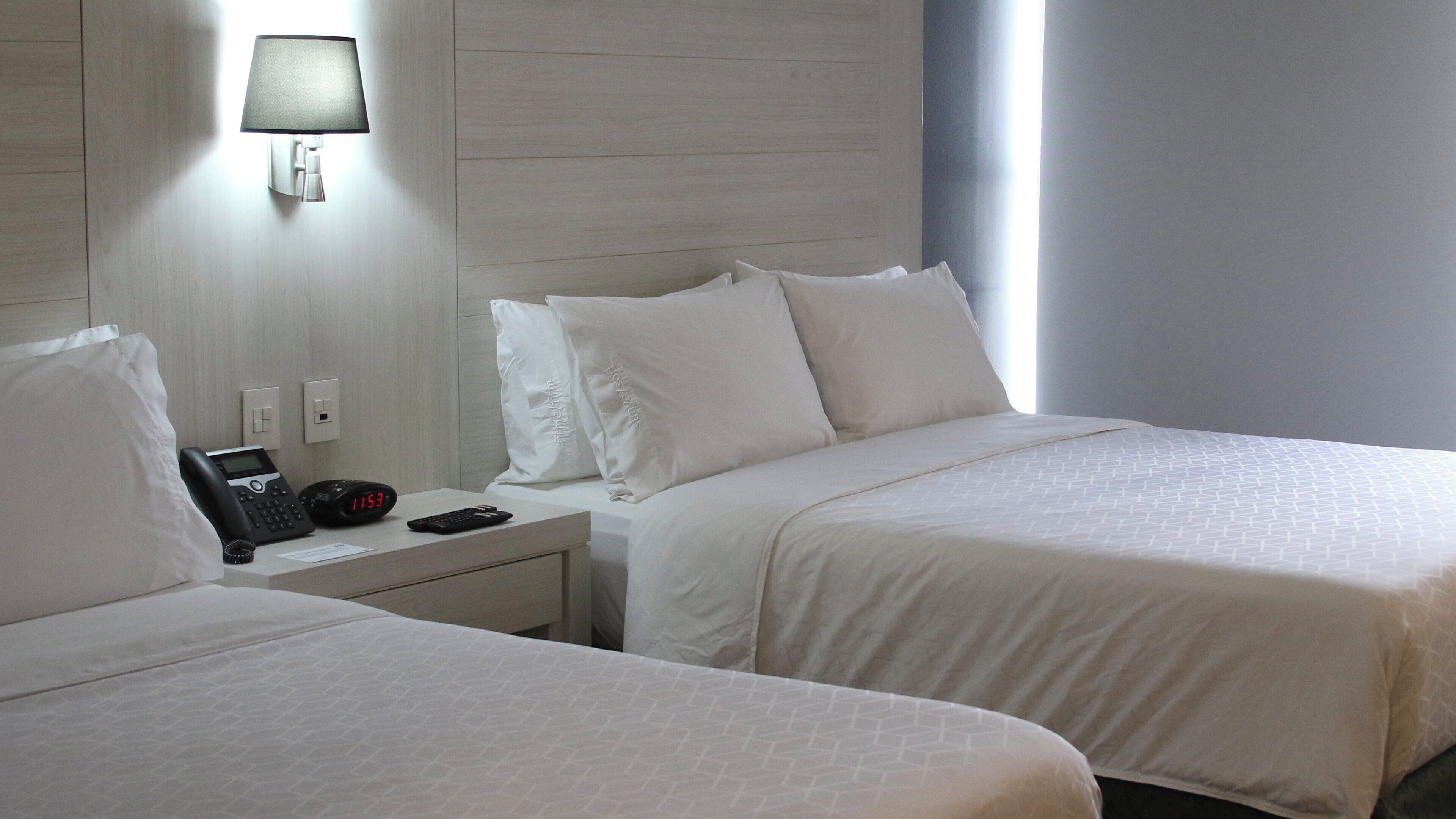 A hotel room with two beds, a bedside table with a phone and alarm clock, a wall-mounted lamp, and a window with natural light.
