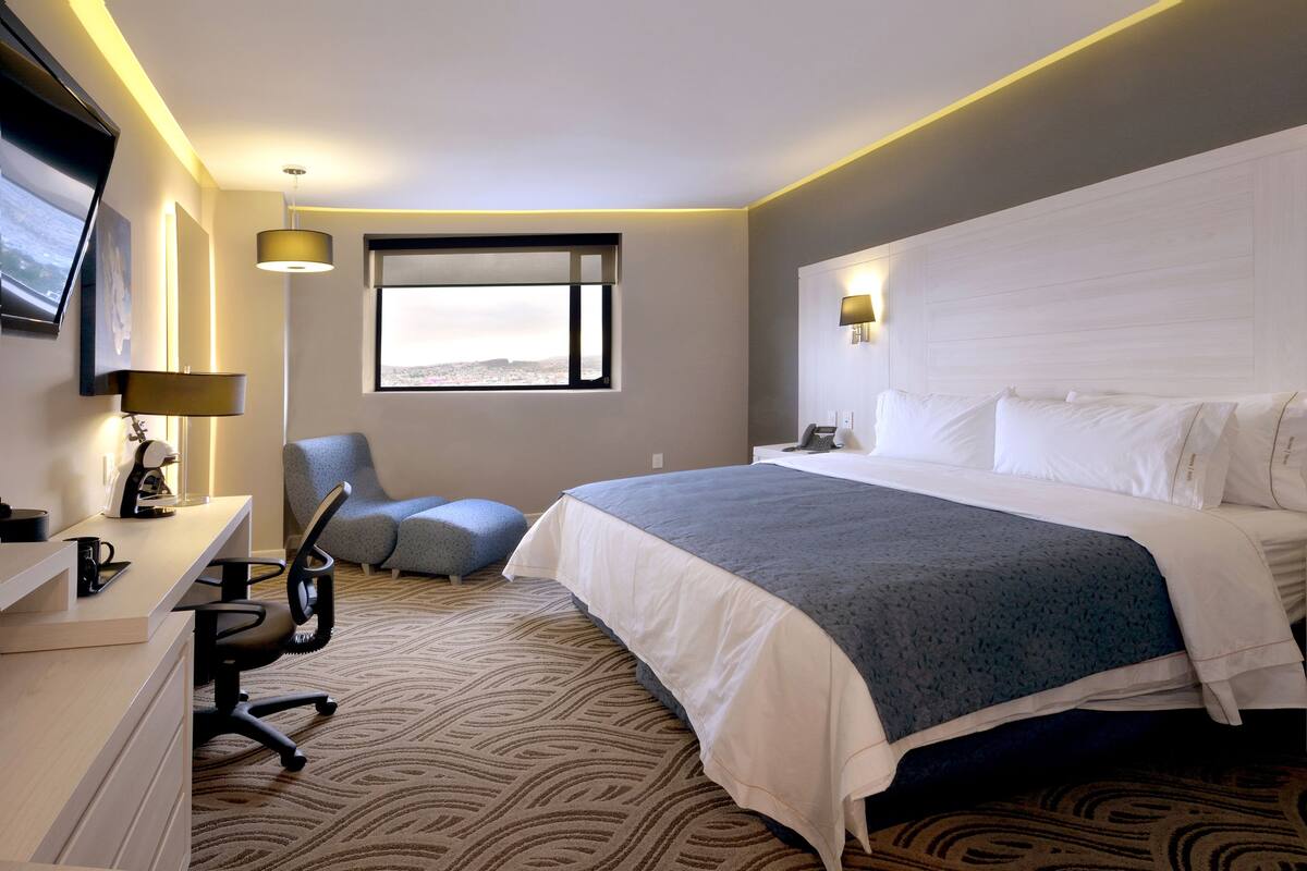 A hotel room with a large bed, a desk with a chair, a desk lamp, a phone, and a window with a view.