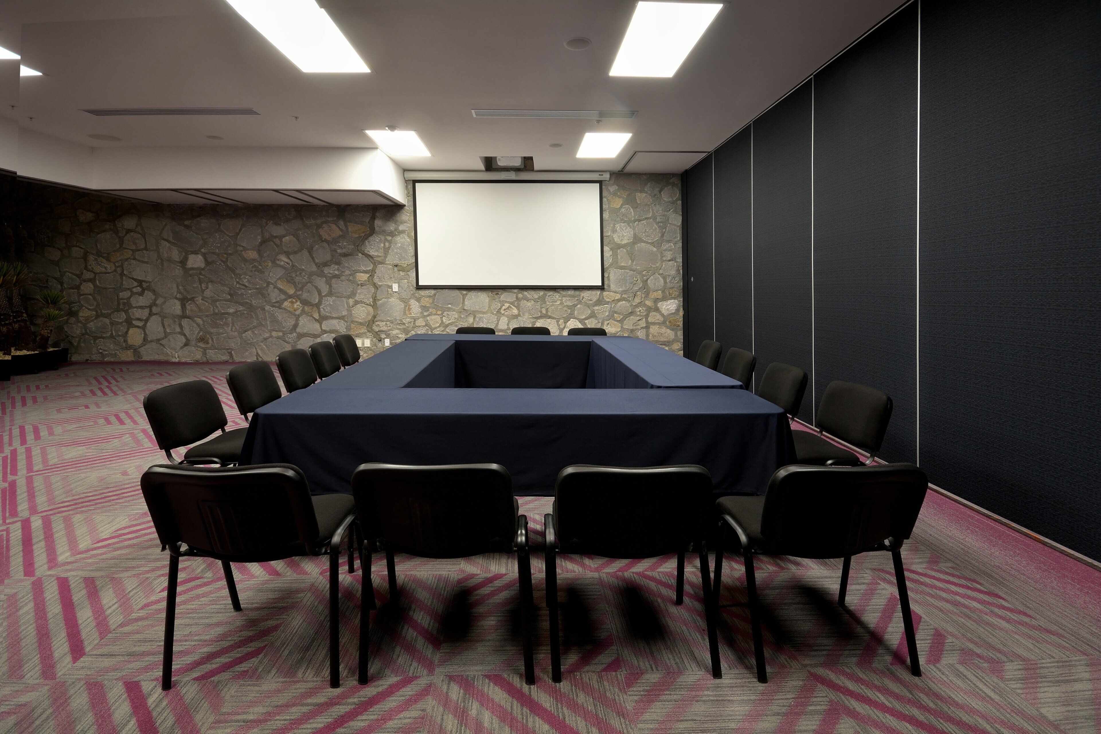 A conference room with a long table, chairs, a projector screen, and a stone wall.