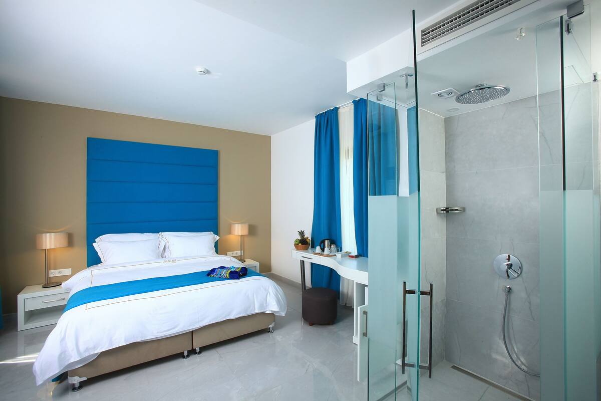 Quarto de hotel moderno com uma cama grande, uma escrivaninha, chuveiro e área de duche com parede de vidro.