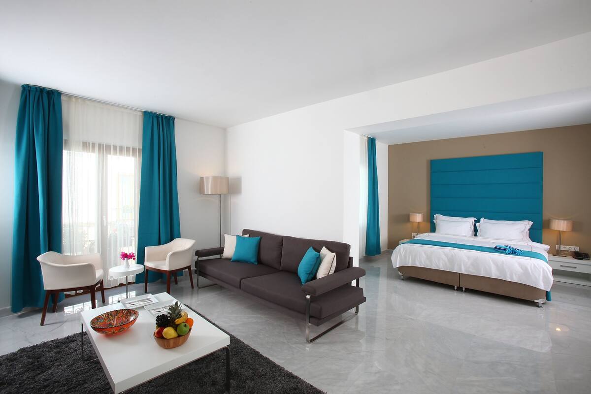 Quarto de hotel moderno com sofá, duas cadeiras, mesa de jantar com frutas, cama com cabeceira azul e mesa de cabeceira com luminárias.