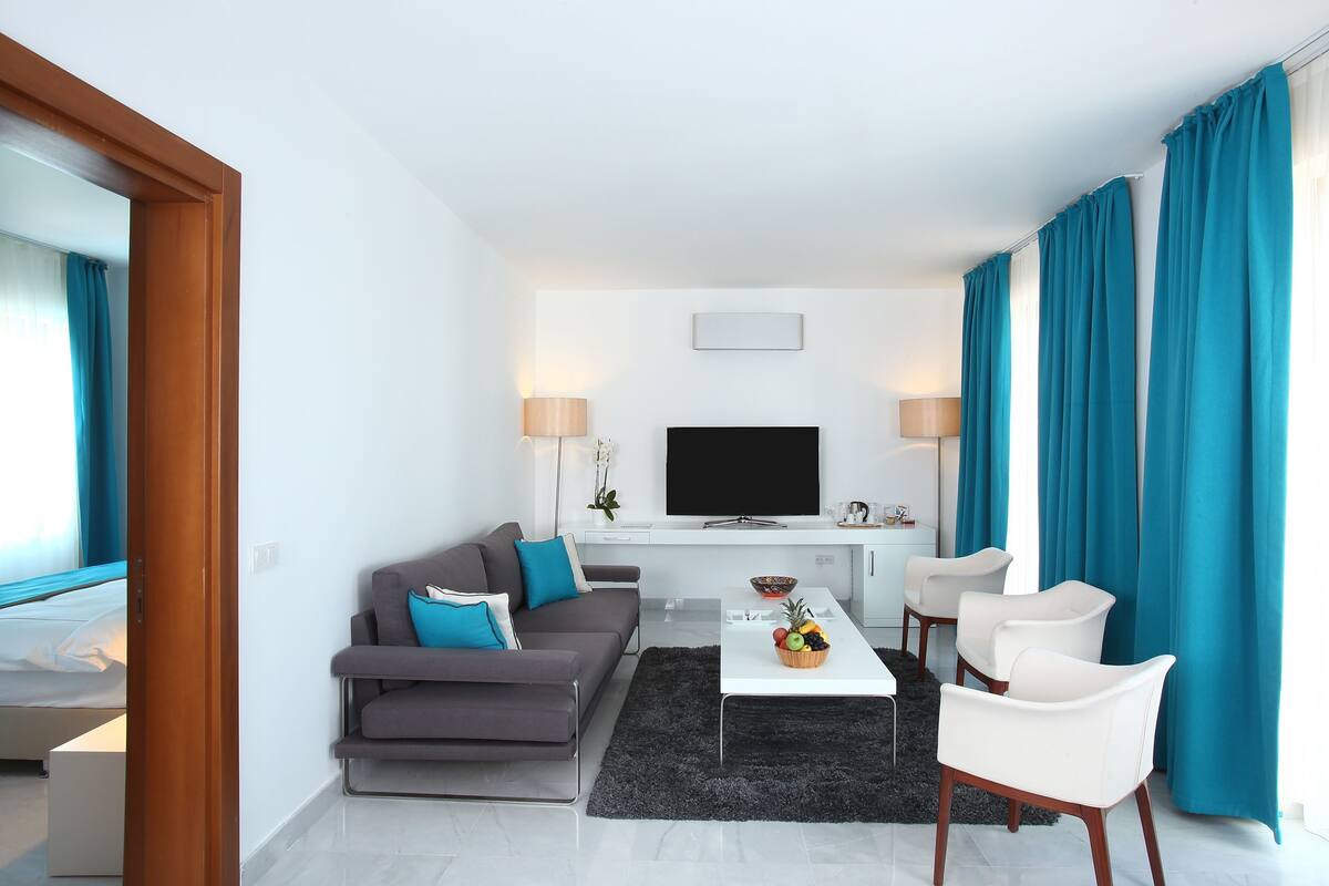 Uma sala de estar moderna com um sofá cinza, poltronas brancas, uma mesa de centro com frutas, uma TV de tela plana e cortinas verde-água.
