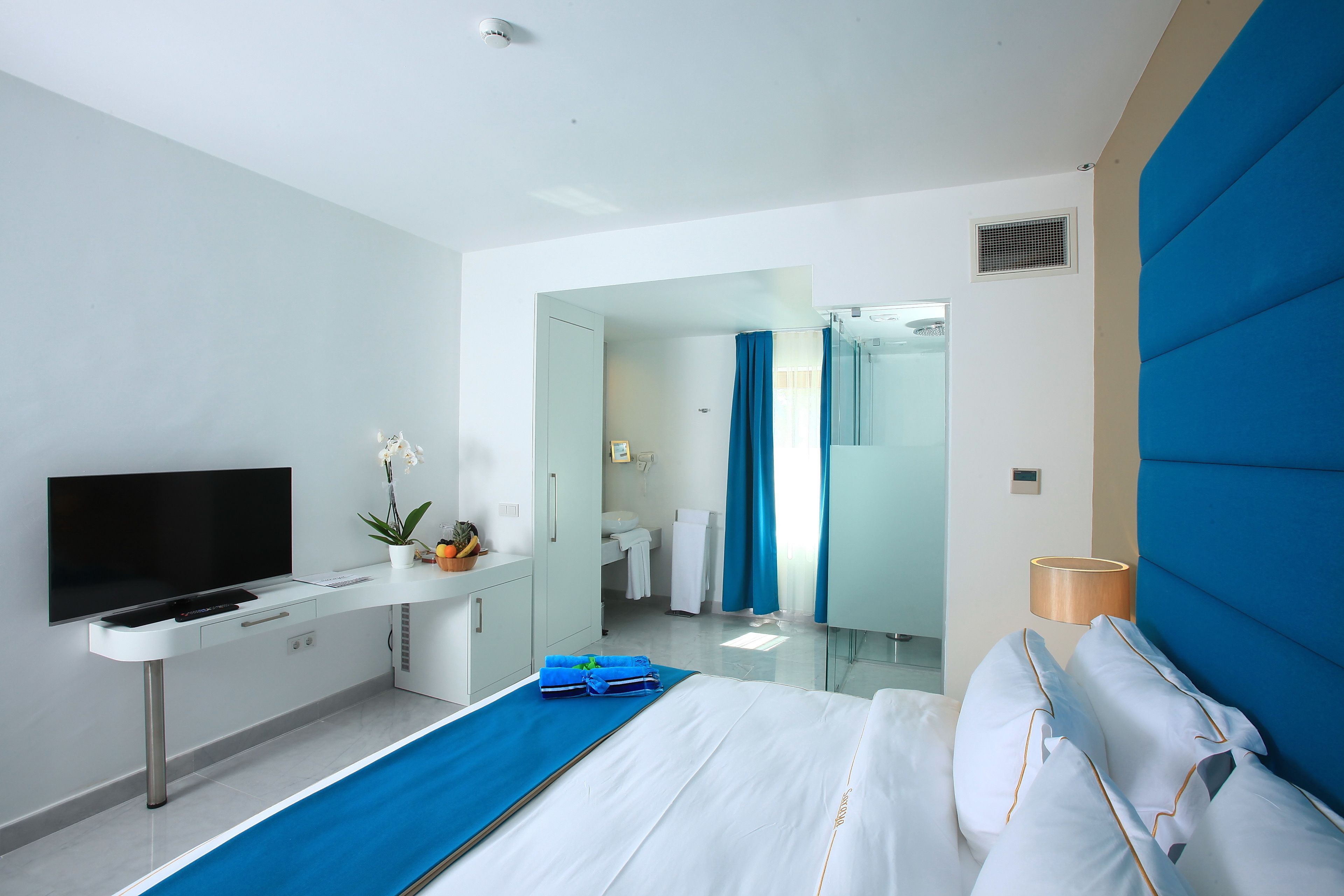 A modern hotel room with a bed, a desk with a flat-screen TV, a bathroom visible through an open door, and a blue accent wall.