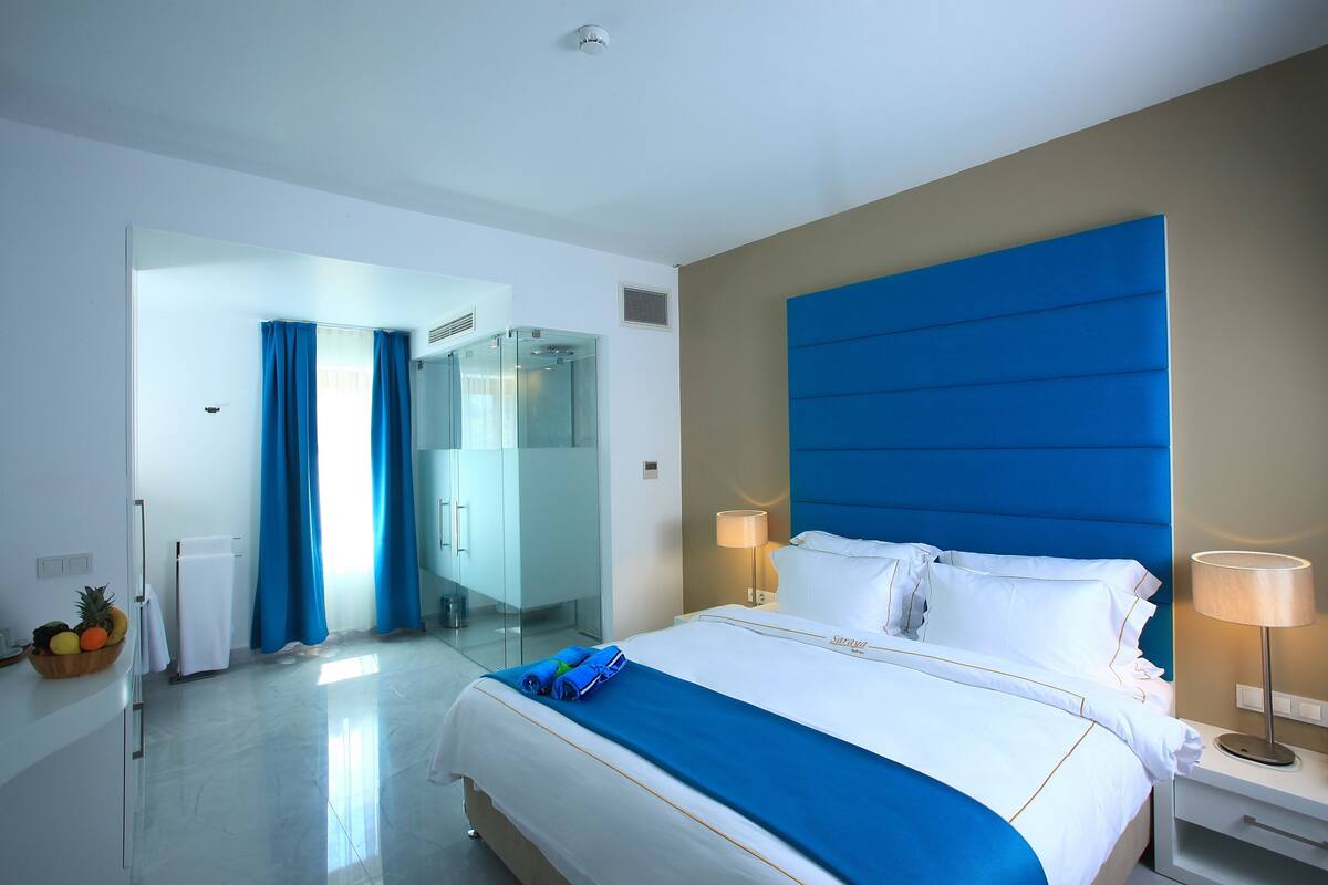 Quarto de hotel moderno com uma cama grande, box de chuveiro de vidro e esquema de cores neutras e claras.