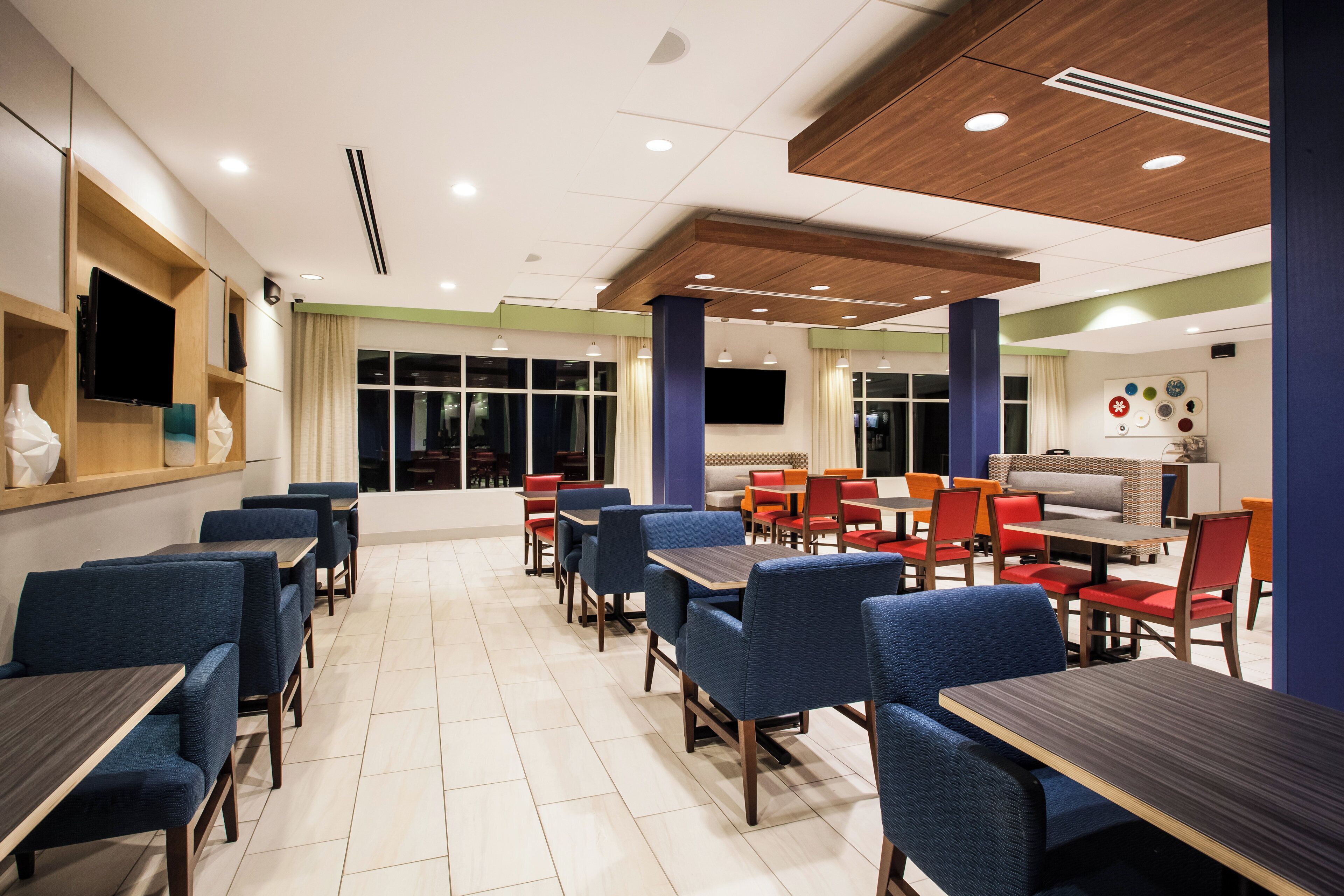 A modern dining area with multiple tables and chairs, a flat-screen TV mounted on the wall, and a ceiling with recessed lighting.