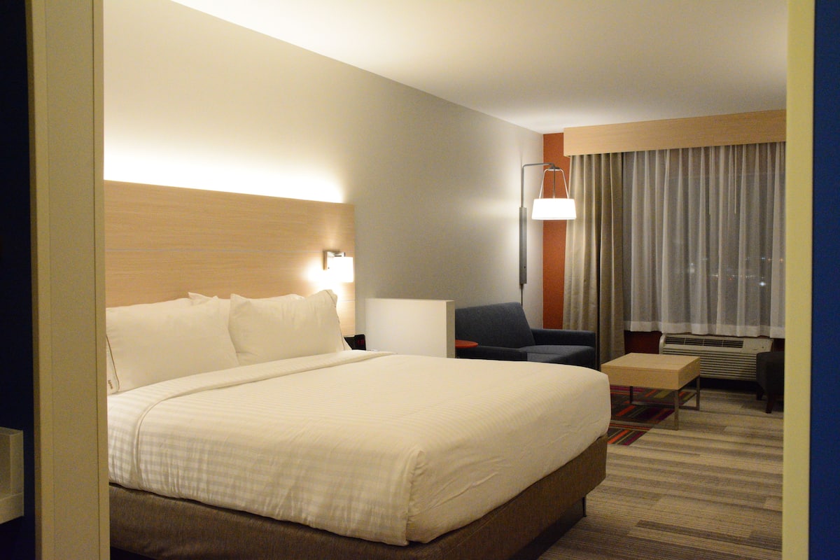 Habitación de hotel con una cama grande, un escritorio, una silla, una lámpara y una ventana con cortinas.