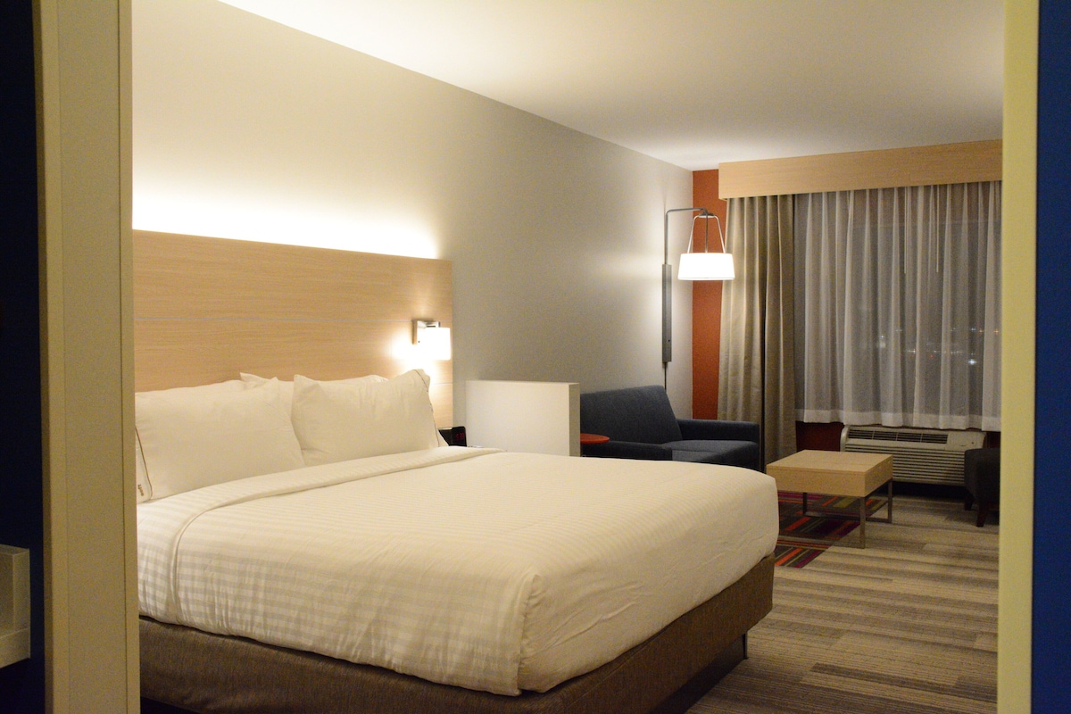 A hotel room with a large bed, a desk, a chair, a lamp, and a view of the city through the window.