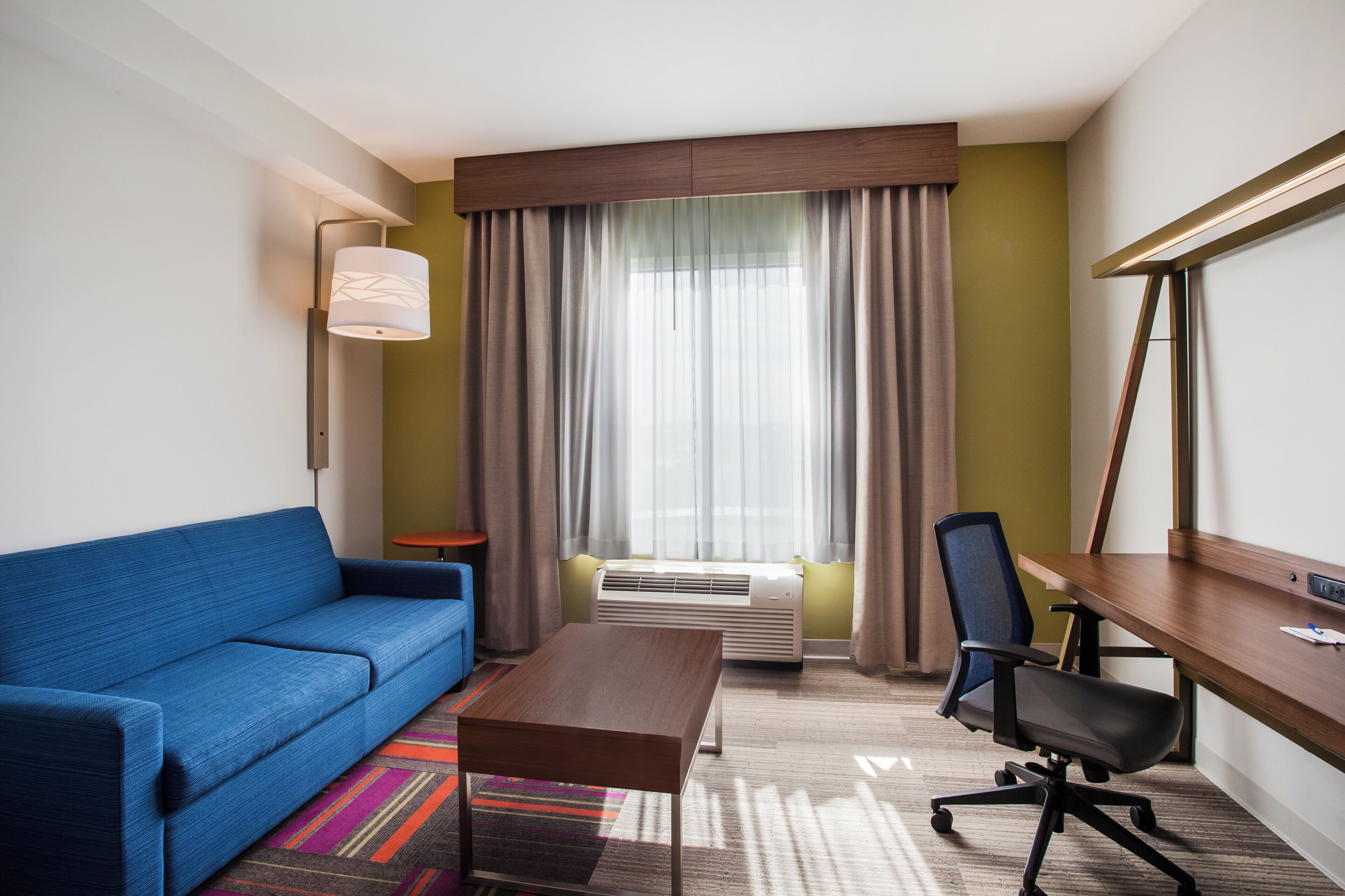A hotel room with a blue sofa, a wooden desk, a chair, a standing lamp, and a large window with curtains.