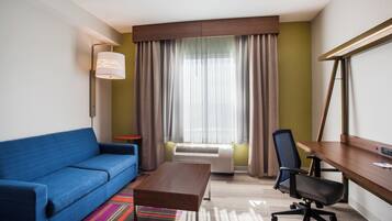 A hotel room with a blue sofa, a wooden desk, a chair, a standing lamp, and a large window with curtains.