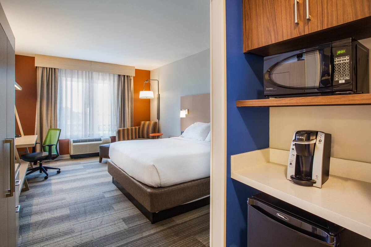 Habitación de hotel con cama, escritorio, silla, aire acondicionado y un microondas en un estante.