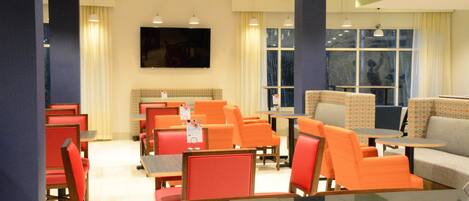 A hotel room with a television, orange and red seating, a large window, and a flat-screen TV mounted on the wall.