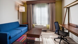 Una habitación de hotel con un sofá azul, un escritorio de madera, una silla de oficina negra, una lámpara de pie y un ventanal con cortinas.