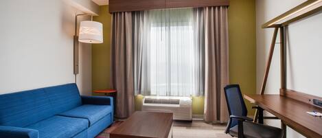 A hotel room with a blue sofa, a wooden desk, a black office chair, a standing lamp, and a large window with curtains.