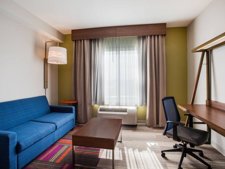 Una habitación de hotel con un sofá azul, un escritorio de madera, una silla de oficina negra, una lámpara de pie y un ventanal con cortinas.