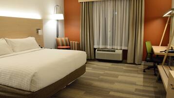 A hotel room with a large bed, a desk, a chair, a lamp, and an air conditioning unit.
