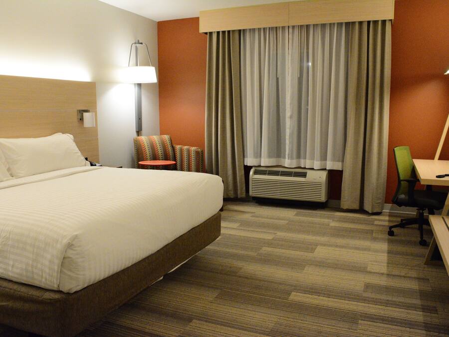 A hotel room with a large bed, a desk, a chair, a lamp, and an air conditioning unit.