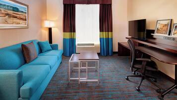 Habitación de hotel con sofá azul, una mesita, un escritorio con computadora y un televisor.