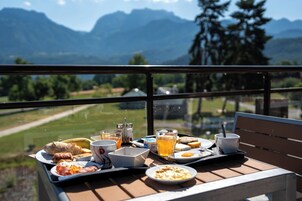 Bandeja de café da manhã com diversos alimentos e bebidas em uma varanda com vista para uma paisagem montanhosa.
