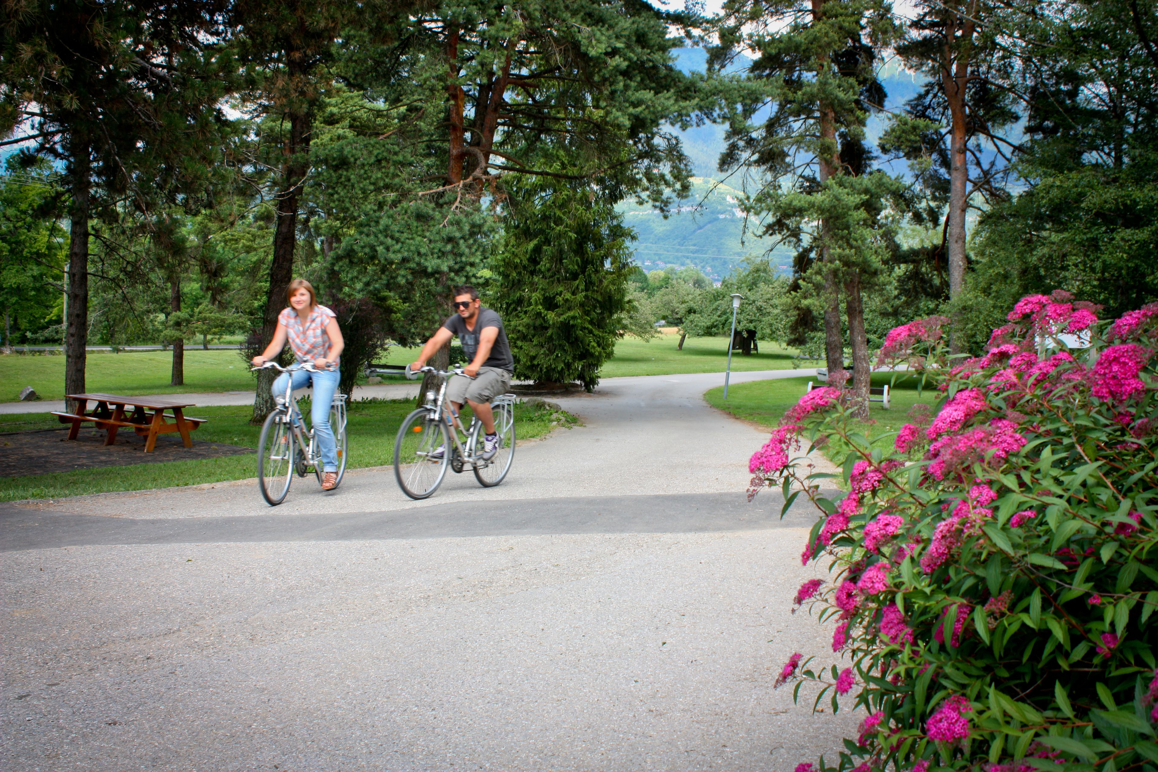 Two people riding bicycles on a paved path in a park with trees and flowers.