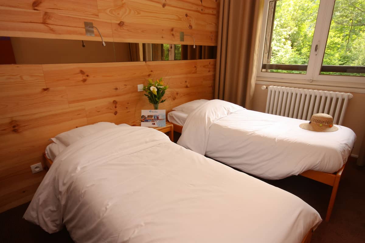 Une chambre avec deux lits, une tête de lit en bois, une table de chevet ornée d’un vase de fleurs et une fenêtre donnant sur des arbres.