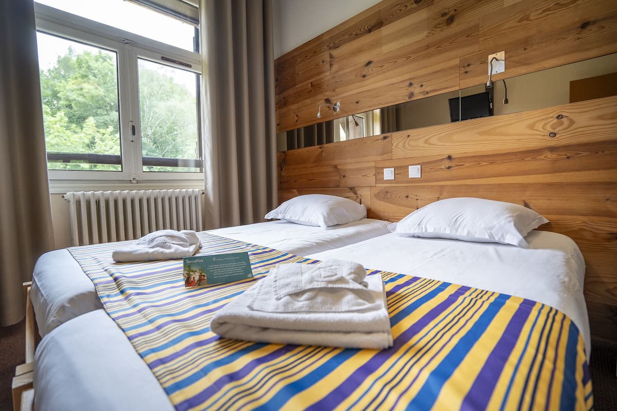 Une chambre d’hôtel avec deux lits, une tête de lit en bois, une fenêtre avec des rideaux et un radiateur.
