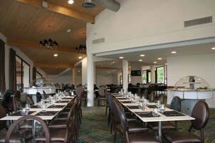 Uma ampla sala de jantar com longas mesas dispostas para vários convidados, destacando-se teto alto com vigas de madeira e iluminação embutida.