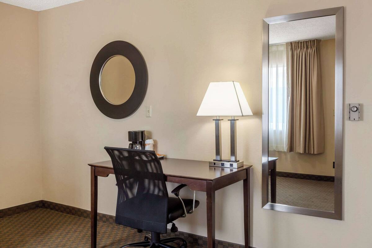 A hotel room with a desk, chair, lamp, and a mirror.