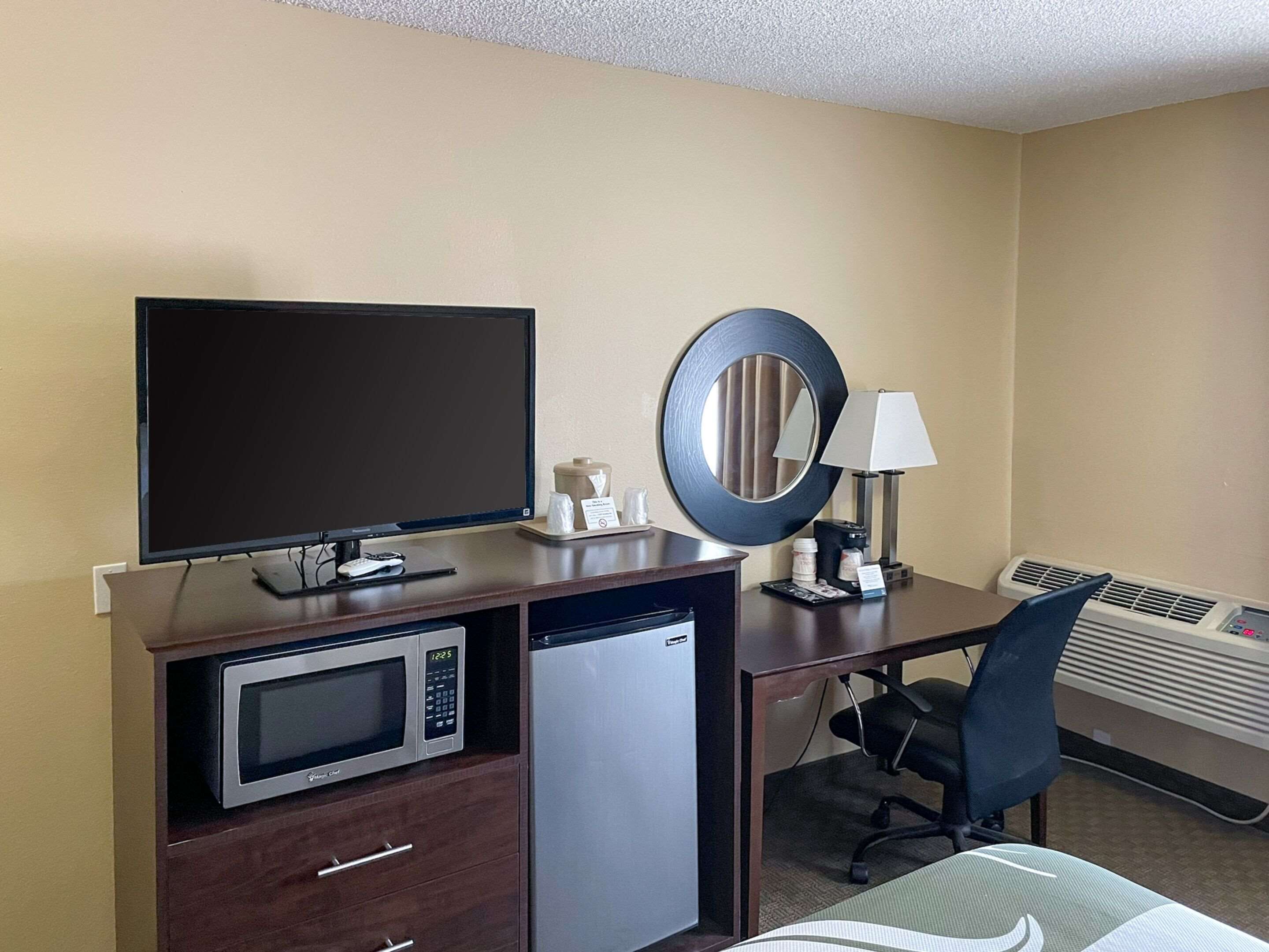 A hotel room with a desk, a television, a microwave, a refrigerator, a mirror, a chair, and an air conditioning unit.