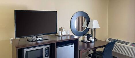 A hotel room with a desk, a television, a microwave, a refrigerator, a mirror, a chair, and an air conditioning unit.