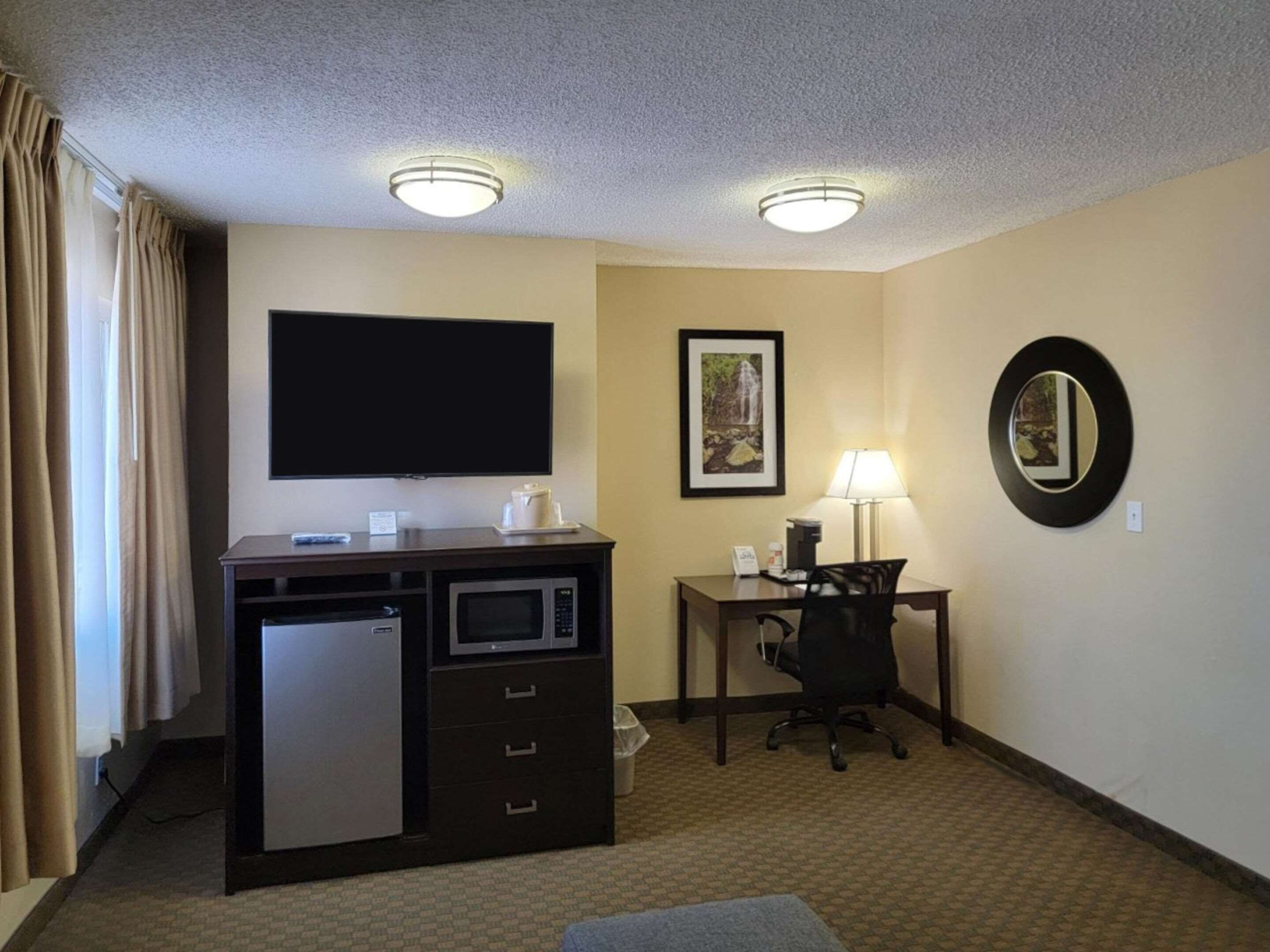 Habitación de hotel con televisión de pantalla plana, escritorio con silla, lámpara, microondas y refrigerador.