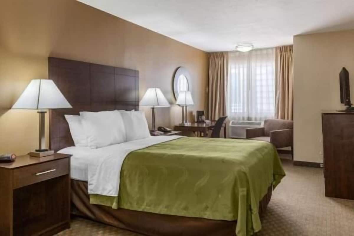 A hotel room with a bed, two bedside lamps, a desk with a chair, a TV, and a window with curtains.