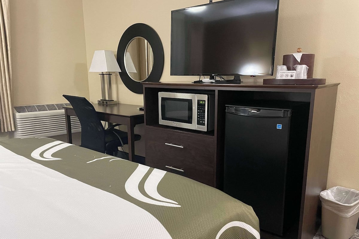 A hotel room with a bed, a desk with a computer, a television, a microwave, a small refrigerator, a trash bin, and a mirror.