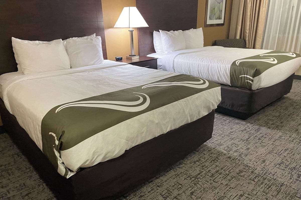 Two double beds with white linens and a green accent, a bedside table with a lamp, and a carpeted floor.
