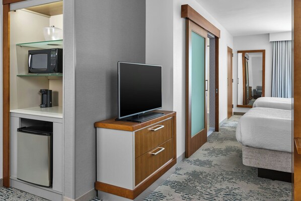 A hotel room with a television on a wooden cabinet, a microwave on a glass shelf, and a refrigerator below.