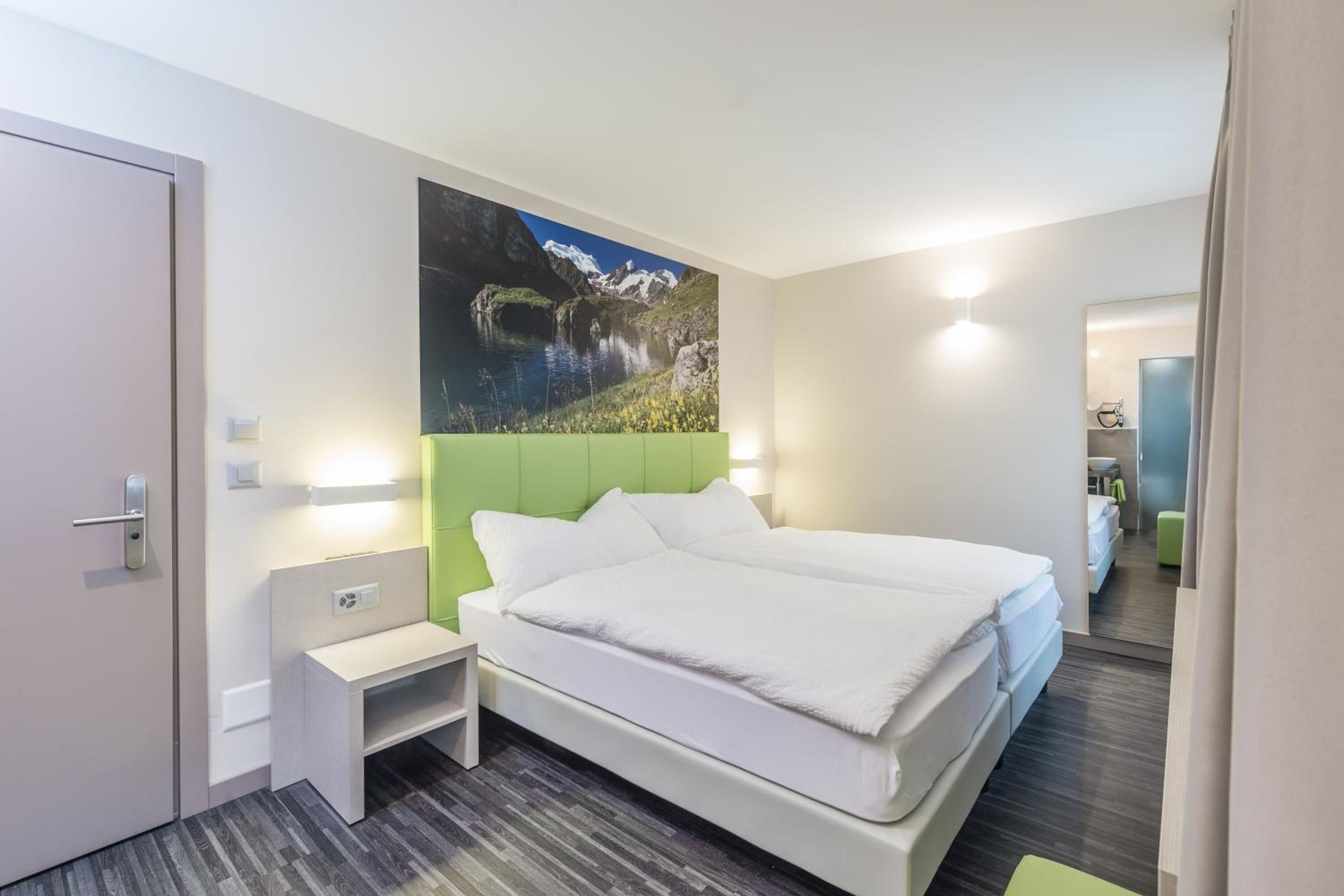 Et moderne hotelværelse med en stor seng, et grønt sengegavl, et natbord og et maleri af en fjeldsø.