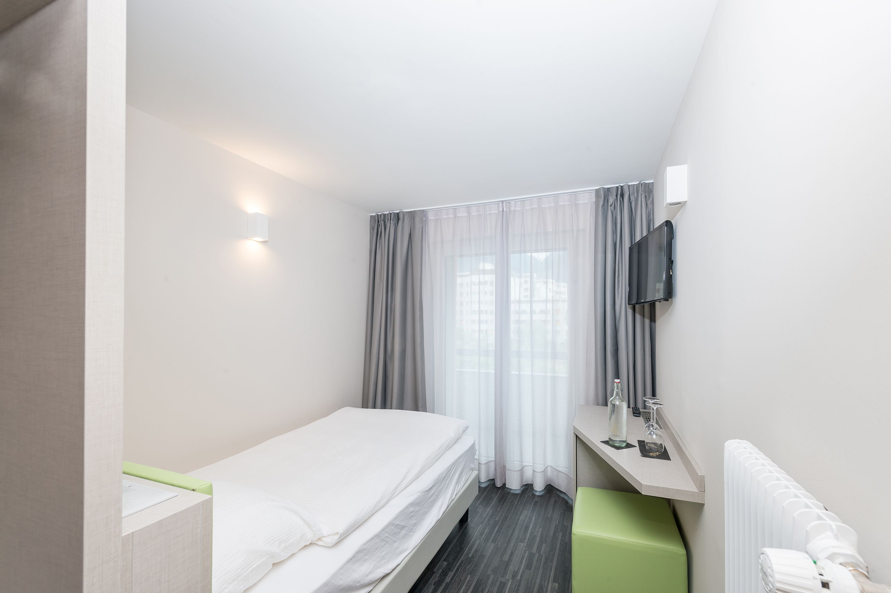 A small, modern hotel room with a single bed, a desk with a chair, a mirror, and a radiator.