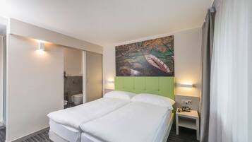A hotel room with a bed, a painting on the wall, a bedside table, and a bathroom visible through a sliding door.
