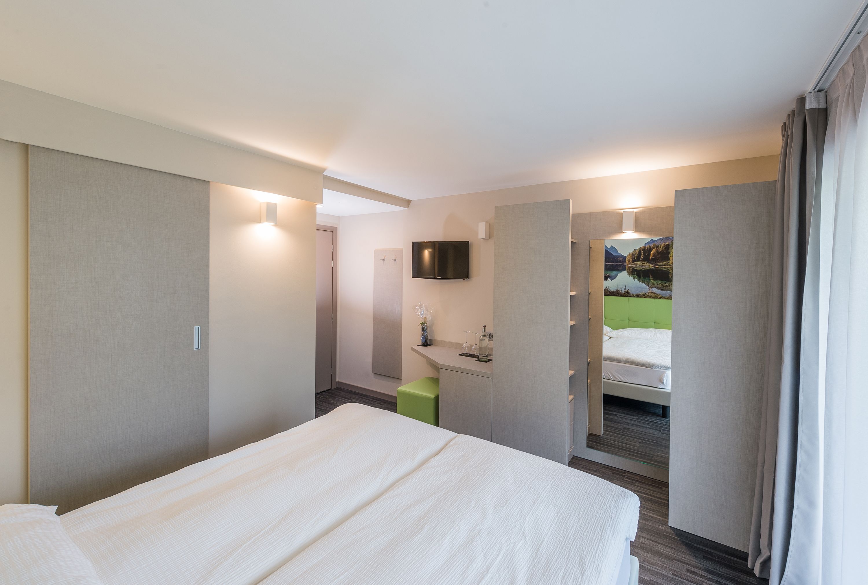 Une chambre d’hôtel avec un lit, un tabouret vert, une télévision, un miroir et une salle de bain avec douche.