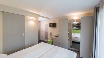 Une chambre d’hôtel avec un lit, un tabouret vert, une télévision, un miroir et une salle de bain avec douche.
