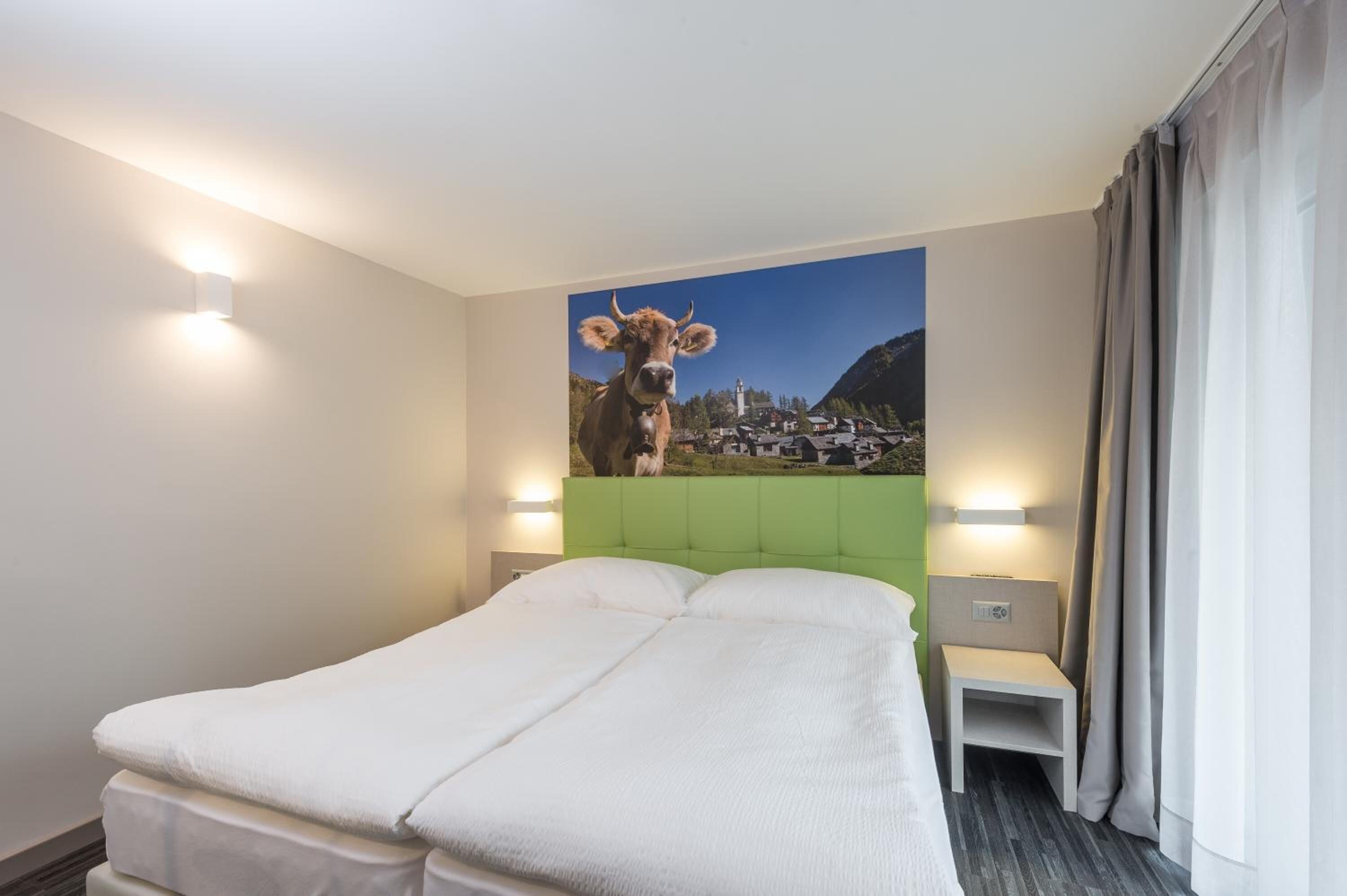 Una habitación de hotel con una cama, cabecera verde, mesita de noche, lámpara y un cuadro con una vaca en la pared.