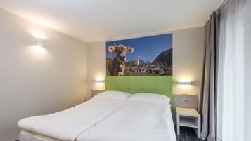 Una habitación de hotel con una cama, cabecera verde, mesita de noche, lámpara y un cuadro con una vaca en la pared.